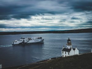 Holburn Head Lighthouse, overlooking a NorthLink ferry sailing across dark waters with a grey cloudy sky in the background